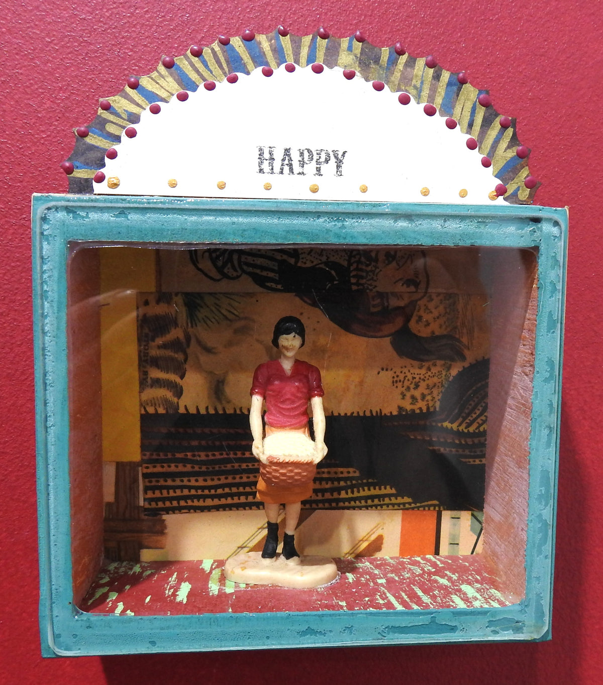 A handcrafted wooden shadow box display mounted against a red wall. The box features a distressed turquoise blue frame, a white arched top panel with the word 'HAPPY' in black, and a colorful collage of vintage posters and images inside.